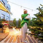 Simple Ways to Keep Your Winter Landscape Looking Beautiful; Woman decorating Christmas tree at beautiful backyard of her counry house. Concept of preparation for the winter holidays. Caucasian woman wearing green sweater and hat. Wide angle view