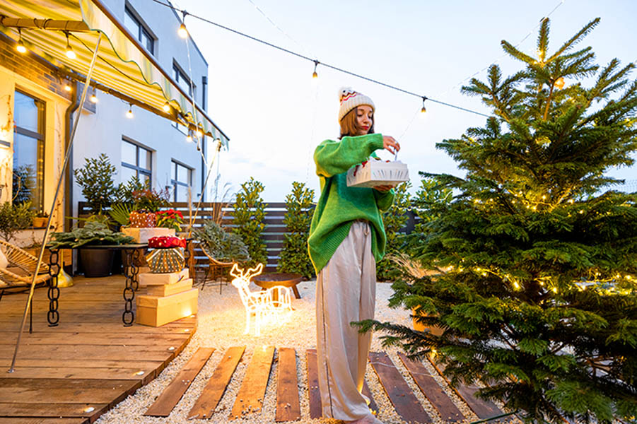 Simple Ways to Keep Your Winter Landscape Looking Beautiful; Woman decorating Christmas tree at beautiful backyard of her counry house. Concept of preparation for the winter holidays. Caucasian woman wearing green sweater and hat. Wide angle view